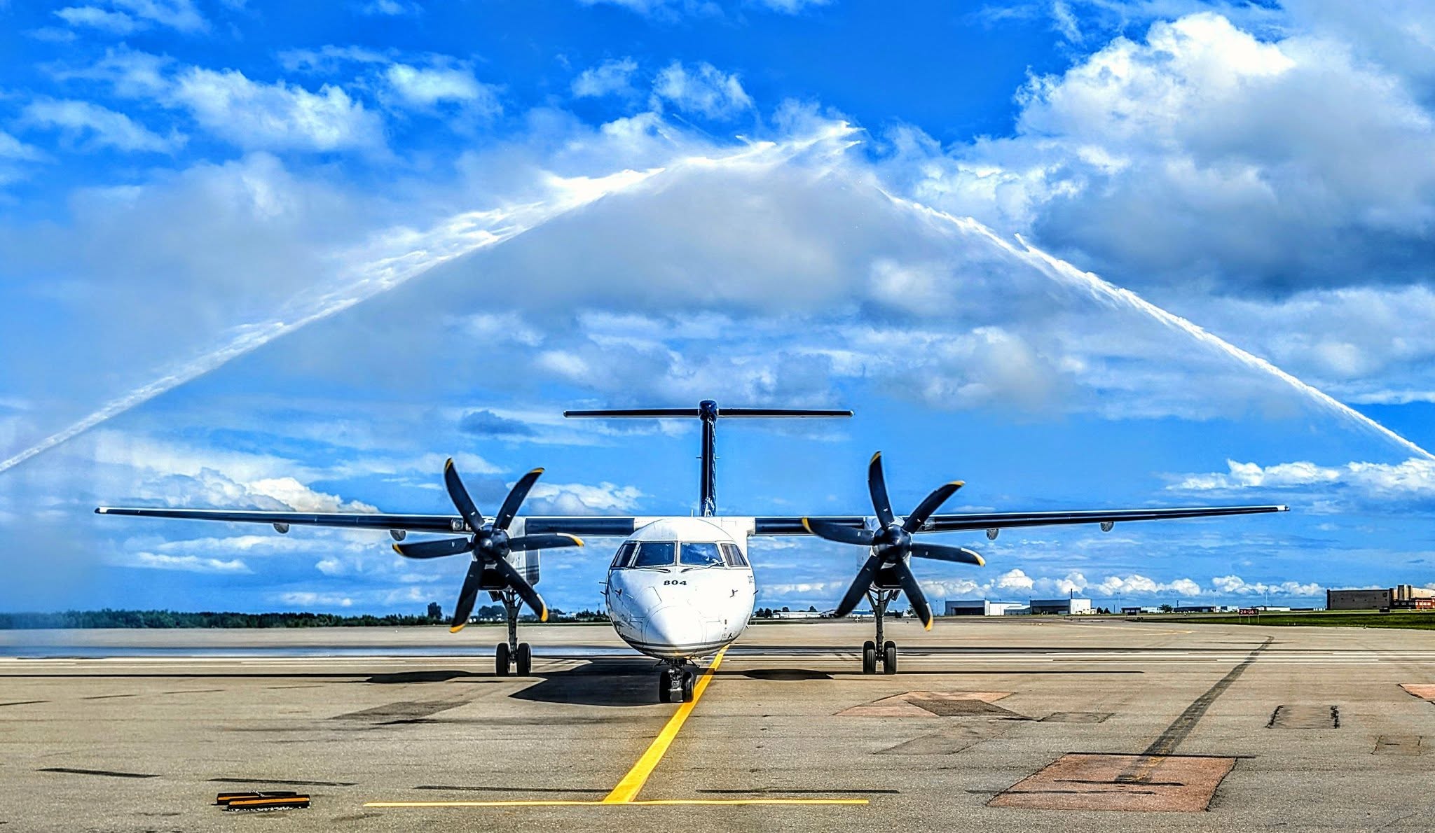 A twin-engine turboprop airplane on an airport tarmac is being honored with a water cannon salute, with streams of water arching over its wings and fuselage against a bright blue sky with fluffy clouds.