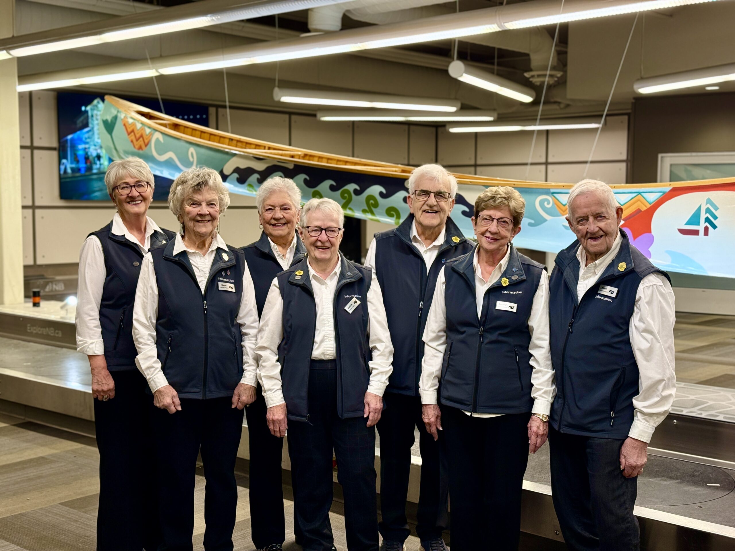 Seven individuals wearing navy blue vests over white shirts stand together indoors, with a decorated canoe suspended overhead in the background.