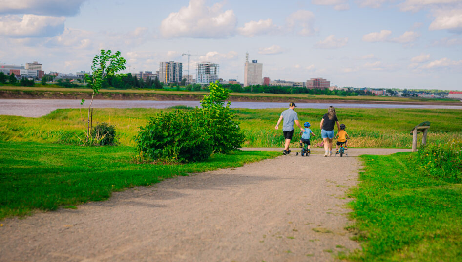 A family walks and bikes along a riverside trail in Moncton, with green marshland, the river and the downtown skyline in the background.