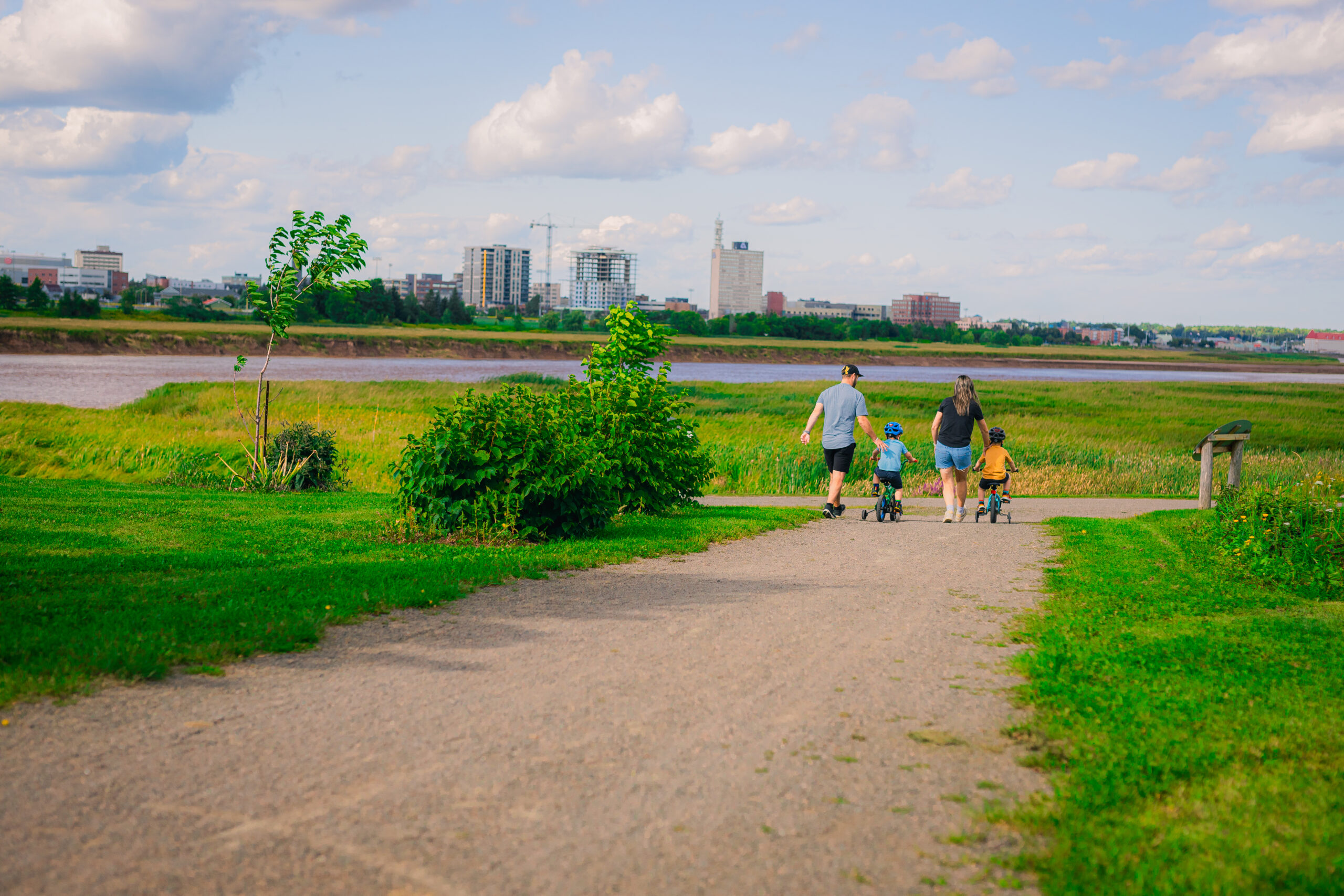 A family walks and bikes along a riverside trail in Moncton, with green marshland, the river and the downtown skyline in the background.