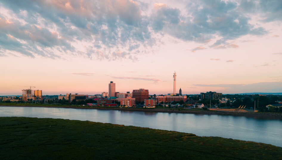 Aerial view of downtown Moncton at sunset, showing city buildings, streets, green spaces and a gathering in a public plaza.
