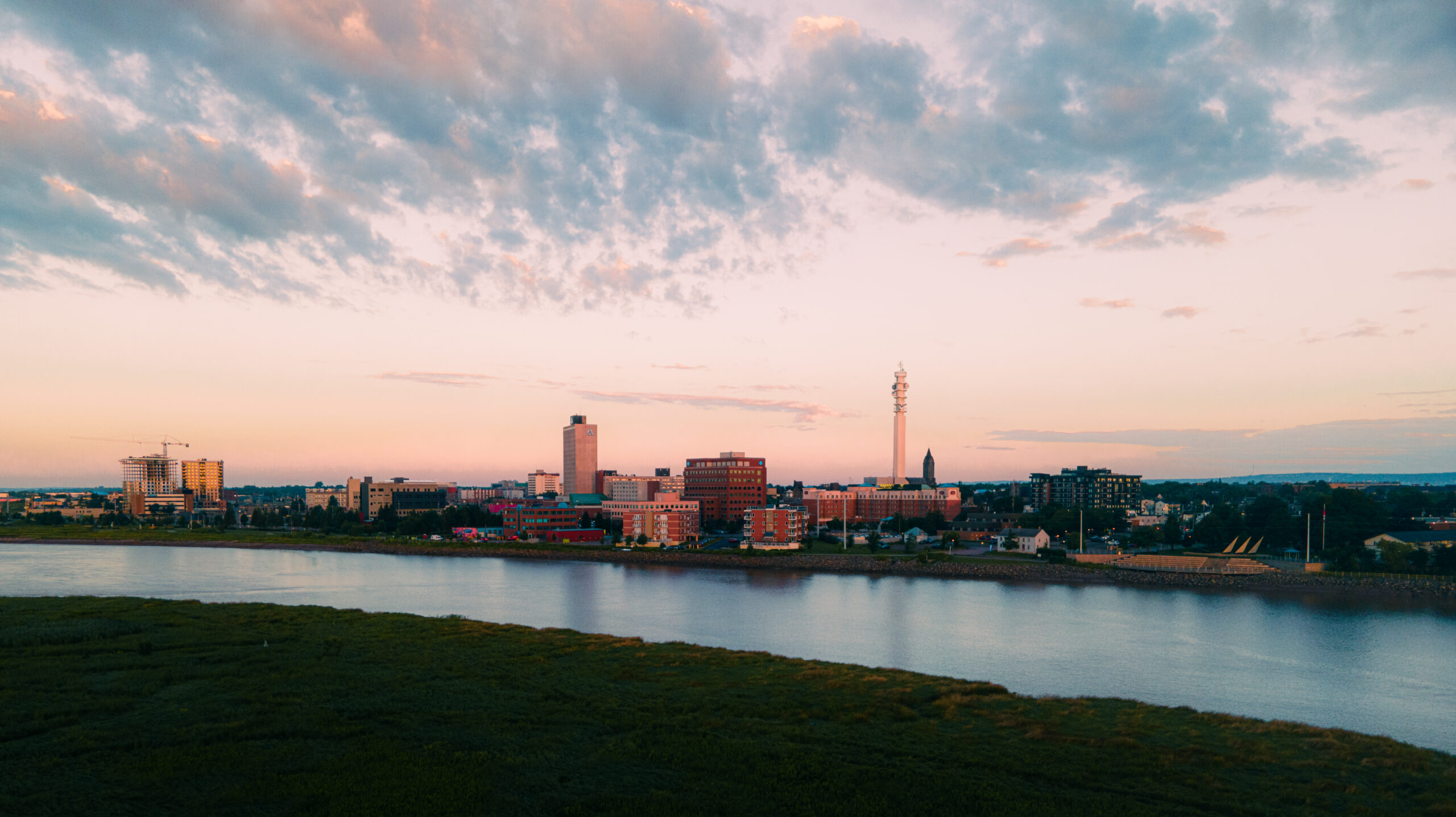 Aerial view of downtown Moncton at sunset, showing city buildings, streets, green spaces and a gathering in a public plaza.